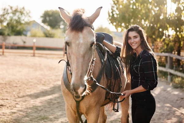 Dziewczyna przygotowuje konia do jazdy na torze wyścigowych. Wesoła kobieta Jockey zapiąć siodełko, uwodzenie cute zwierząt i uśmiechnięta radosna kamera, cieszyć się świeżym powietrzem, biorąc Rancho opieki, zwierzęta gospodarskie — Zdjęcie stockowe