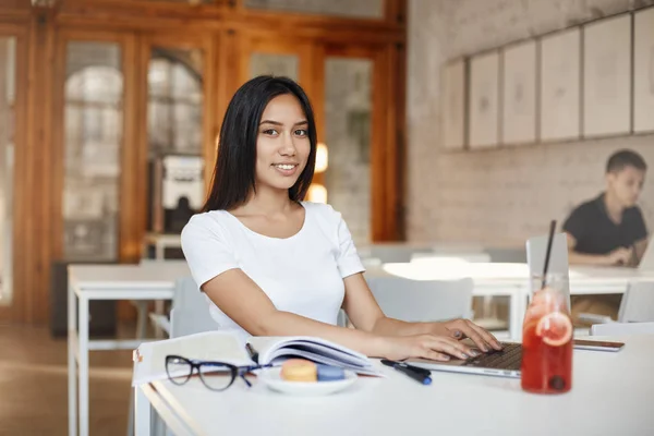 Sonriendo hermosa estudiante joven que estudia en la cafetería, la cámara sonriente como ensayo de mecanografía utilizando el ordenador portátil, rodeado de libros, deberes y vasos, beber jugo para ganar energía, concentrar el trabajo —  Fotos de Stock