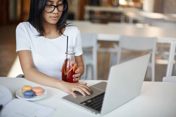 Ritagliato pensoso e concentrato intelligente studentessa in occhiali studiando in caffè, navigare in internet in computer portatile, tenere bottiglia di vetro di succo, fissare display del computer focalizzato, imparare online — Foto Stock