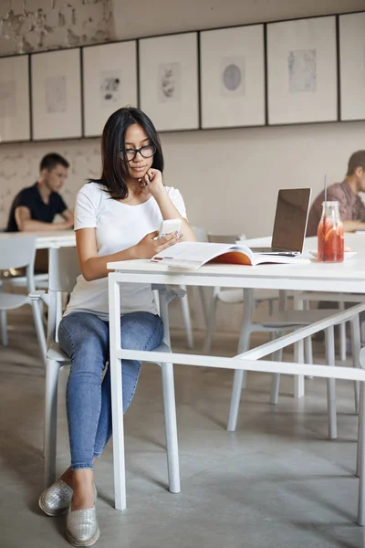 Vertical disparo ocupada joven mujer en gafas sentarse sola cafetería rodeada de material de trabajo portátil y portátil, utilizando el teléfono inteligente, desplazándose alimentación de las redes sociales durante el descanso de estudiar —  Fotos de Stock