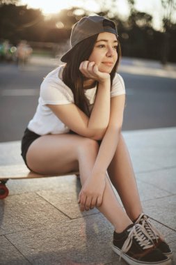 Dikey çekim gri snapback, spor ayakkabı, el eğik kafa ve mutlu gülümseyen rüya gibi kaygısız genç patenci kız, akşam gün batımı sırasında diğer patenciler ile sohbet olarak ahşap kuruş tahtası üzerinde oturup