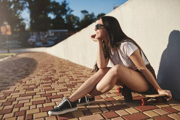 Ungdoms-, fritids-och frilufts koncept. Glad drömmande hipster skater flicka sitta på trä Penny Board nära vita muren titta på sommaren solnedgång, leende glad, lär dig hur utföra Kickflip — Stockfoto