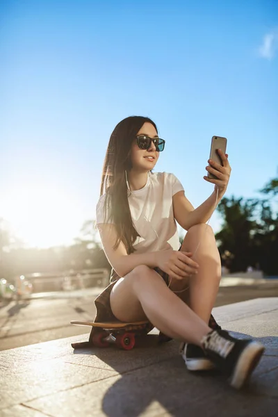 Вертикальный снимок низкого угла обзора стильная, популярная девушка-хипстер в прямом эфире со смартфоном подключается к Wi-Fi, как сидят на деревянной пенни доске, практикуют трюки скейт-парка, слушают музыку проводные наушники — стоковое фото