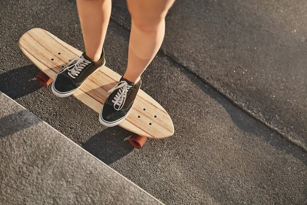 Øvre vinkel med avklipte skudd, hvite hender i joggesko som står på et trebord, gjør kickflip, lær hvordan man gjør triks og stunt på en konsertskatepark med rullebrett, solstråler – stockfoto