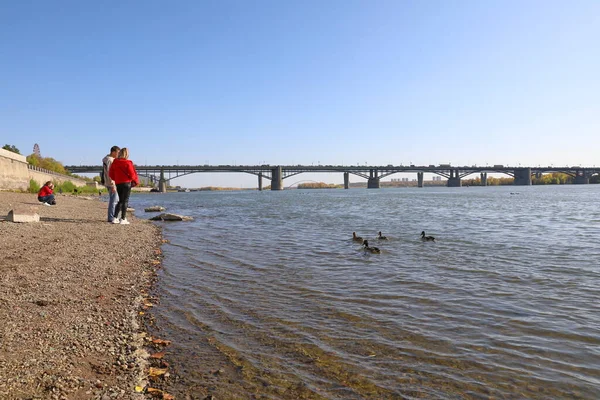 Novosibirsk, RUSSIA-20 Eylül 2020: Oktyabrsky köprüsünün arka planına karşı Ob Nehri kıyısında genç bir adam ve bir kız