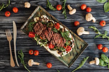 top view of delicious roasted steak, fork with knife and vegetables on wooden table 