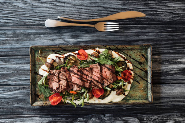 top view of gourmet sliced New York steak, vegetables and fork with knife on wooden table 