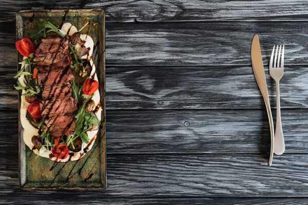 top view of delicious roasted steak with vegetables and fork with knife on wooden table 