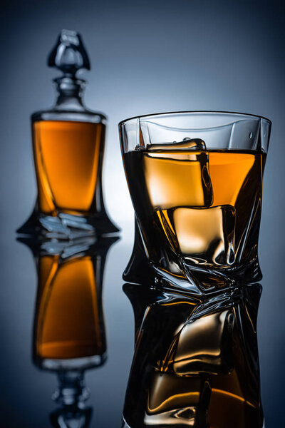selective focus of bottle and glass with whiskey and ice cubes, on grey