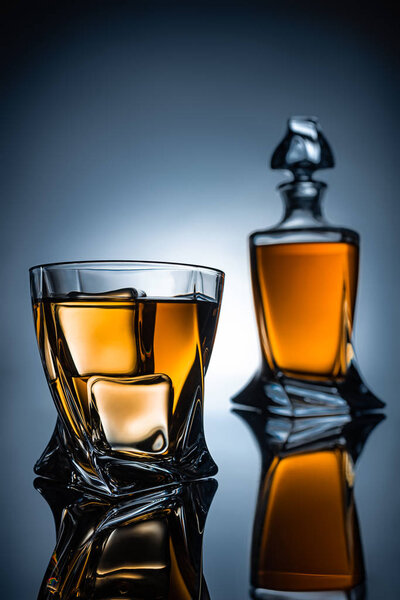 selective focus of whiskey glass with ice cubes and bottle, on grey