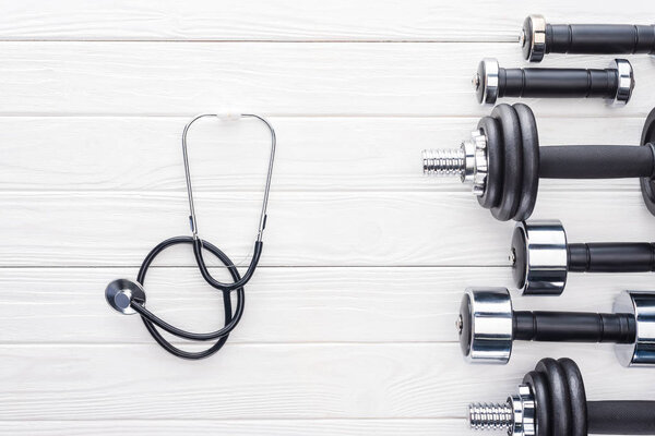 top view of various dumbbells and stethoscope on white wooden surface
