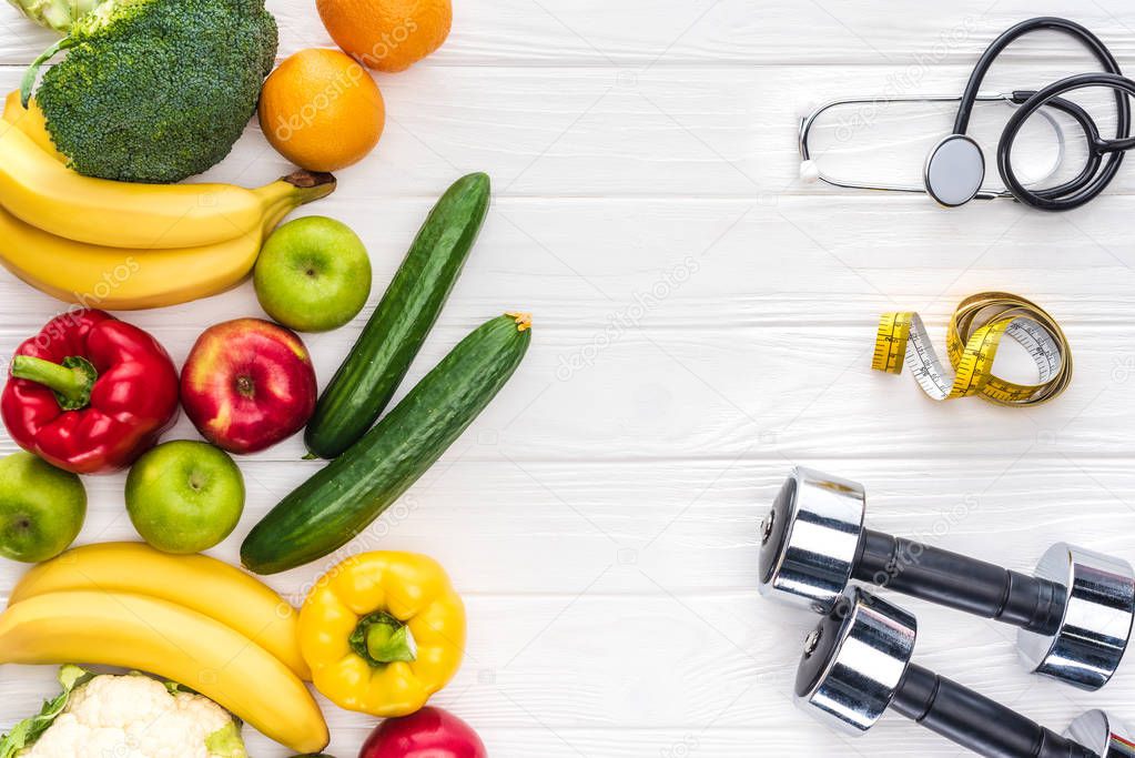 top view of fresh fruits and vegetables, dumbbells, stethoscope and measuring tape on wooden tabletop