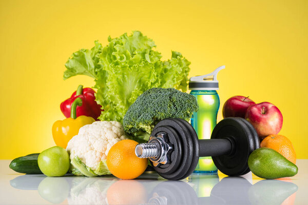 close-up view of dumbbell, bottle of water and fresh fruits and vegetables on yellow 