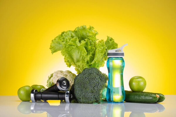 close-up view of fresh green apples and vegetables, dumbbells and bottle of water on yellow