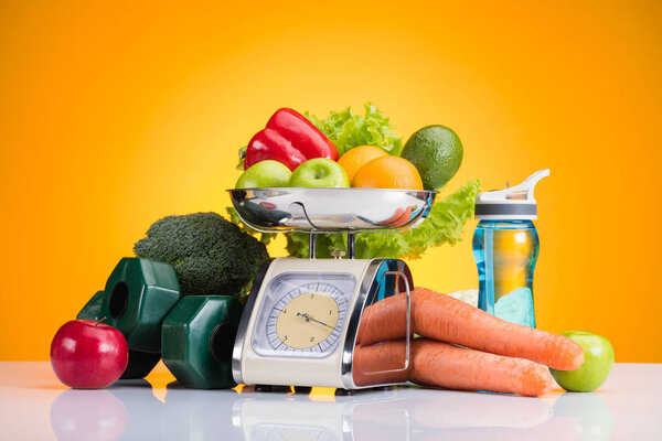 close-up view of fresh fruits and vegetables on scales, dumbbells and bottle of water on yellow 