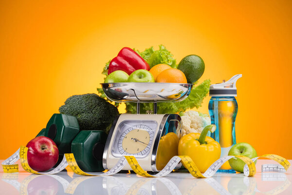 close-up view of fresh fruits and vegetables on scales, sports bottle with water, dumbbells and measuring tape on yellow  