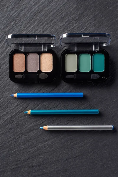 Eye shadows palettes and blue eye pencils on dark slate background