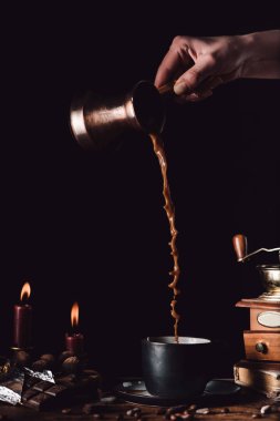 cropped shot of woman pouring coffee from turk into cup at table with chocolate, truffles, candles and coffee grains on black background clipart