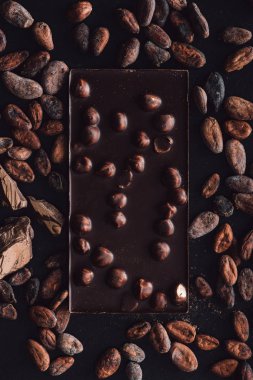 top view of chocolate bar with hazelnuts surrounded by cocoa beans on dark surface clipart
