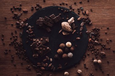 elevated view of cutting board with various types of chocolate pieces and truffles surrounded by cocoa beans, coffee grains and nutmegs on wooden table clipart