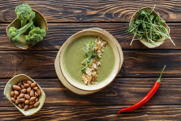 top view of arranged vegetarian cream soup, sprouts, almonds, fresh broccoli in bowls and chili peppers on wooden surface