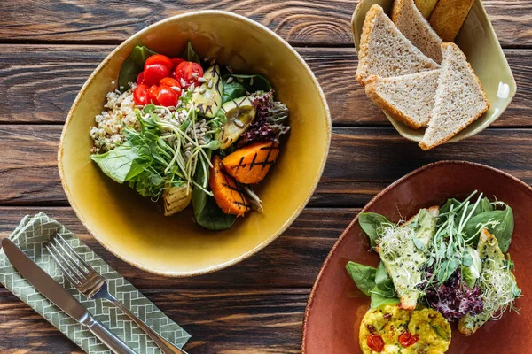 flat lay with vegetarian salads served in bowls, pieces of bread and cutlery on wooden tabletop