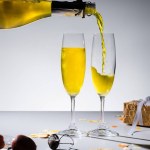 Close up view of pouring yellow champagne into glassware process and arranged gifts on grey backdrop