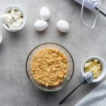 Flat lay with crushed cookies, raw chicken eggs and butter for making pie on grey surface