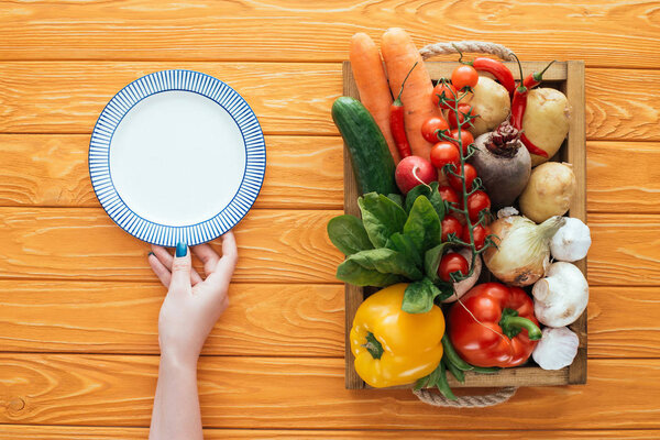 cropped shot of person holding empty round plate and fresh raw vegetables in box on wooden table top