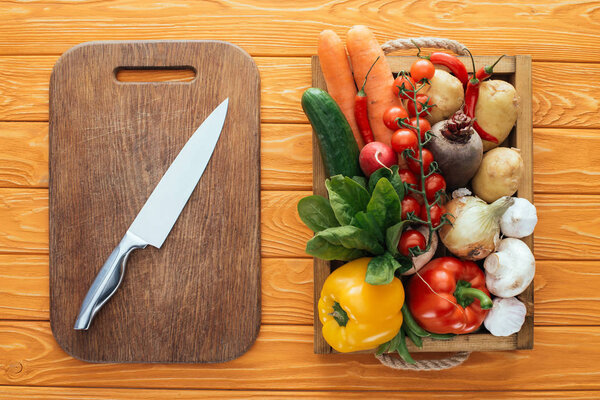 top view of wooden cutting board with knife and fresh raw vegetables in box on table top