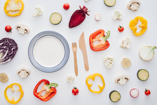 top view of empty round plate, wooden fork with knife and fresh sliced vegetables isolated on white