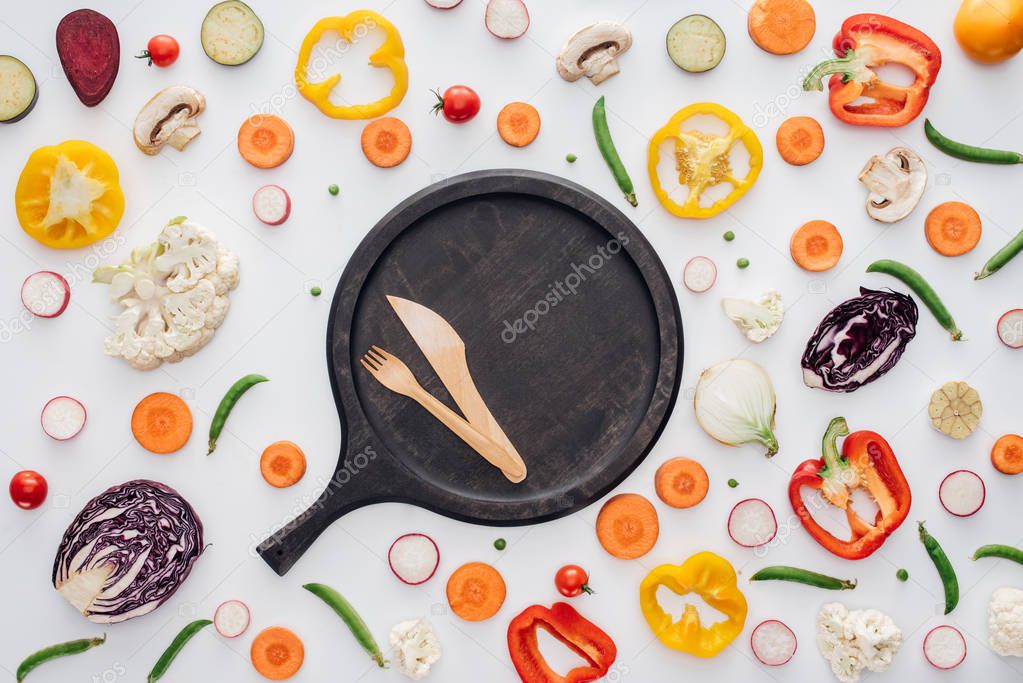 top view of fork and knife on round wooden board and fresh sliced vegetables isolated on white 