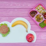 Vista superior de la bandeja con almuerzo para niños para la escuela, hamburguesa y frutas en la mesa rosa