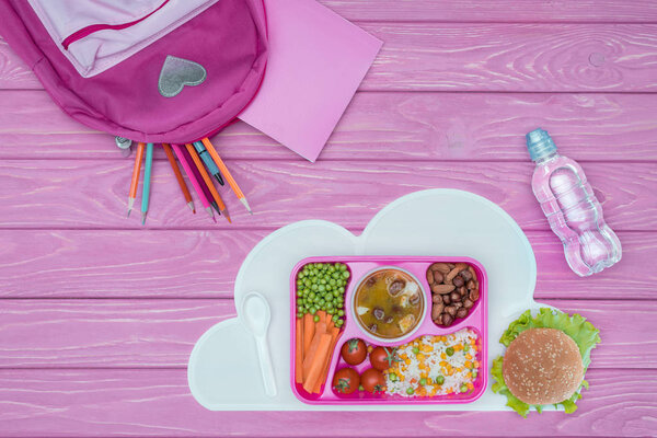 top view of tray with kids lunch for school, pink bag and pencils on pink table