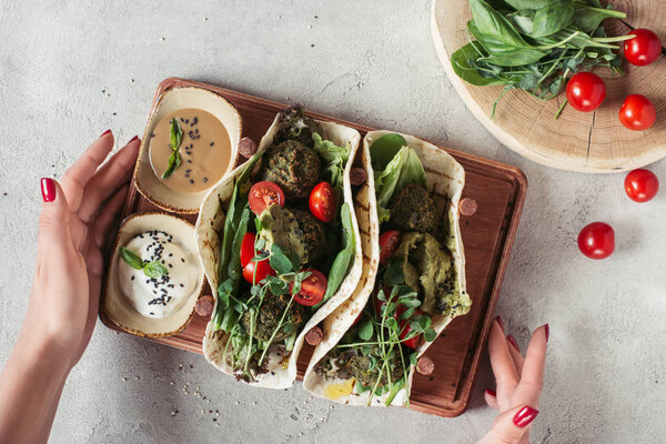 cropped shot of woman holding falafel with tortillas, cherry tomatoes and germinated seeds of sunflower served on wooden board on grey surface