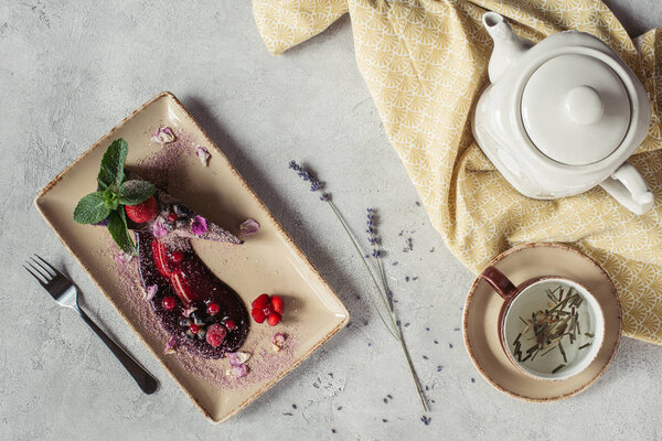 food composition with piece of blueberry cake served with mint leaves and violet petals on plate, teapot and cup of herbal tea on grey tabletop