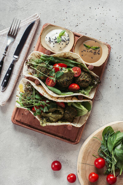top view of falafel with tortillas, cherry tomatoes and germinated seeds of sunflower served on wooden board on grey surface