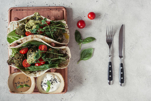 top view of falafel with tortillas, cherry tomatoes and germinated seeds of sunflower served on wooden board on grey surface