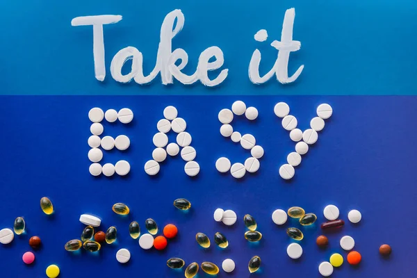 top view of lettering take it easy by white pills near various tablets on blue background