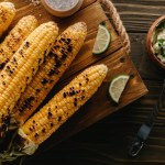 Vue du dessus de la planche à découper avec maïs grillé, tranches de lime près de la brosse de cuisson et beurre avec persil sur table en bois