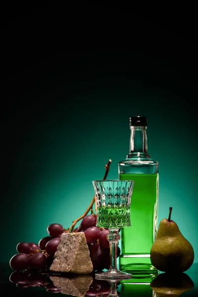 close-up shot of absinthe with fresh pear, grapes and cheese on mirror surface on dark blue background