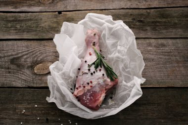 uncooked turkey leg with pepper corns and rosemary on baking paper on wooden tabletop clipart