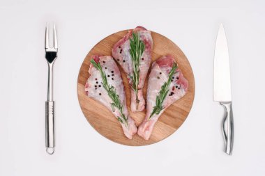 top view of raw turkey legs with pepper corns and rosemary on cutting board with fork and knife, isolated on white clipart