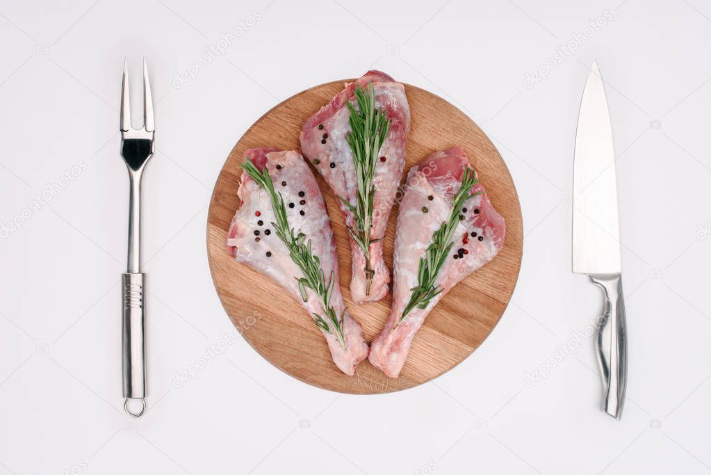 top view of raw turkey legs with pepper corns and rosemary on cutting board with fork and knife, isolated on white