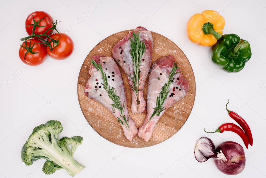 top view of uncooked chicken legs with rosemary on cutting board with raw vegetables near, isolated on white