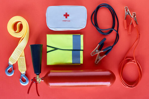 top view of fire extinguisher, first aid kit and jump start cables on red background