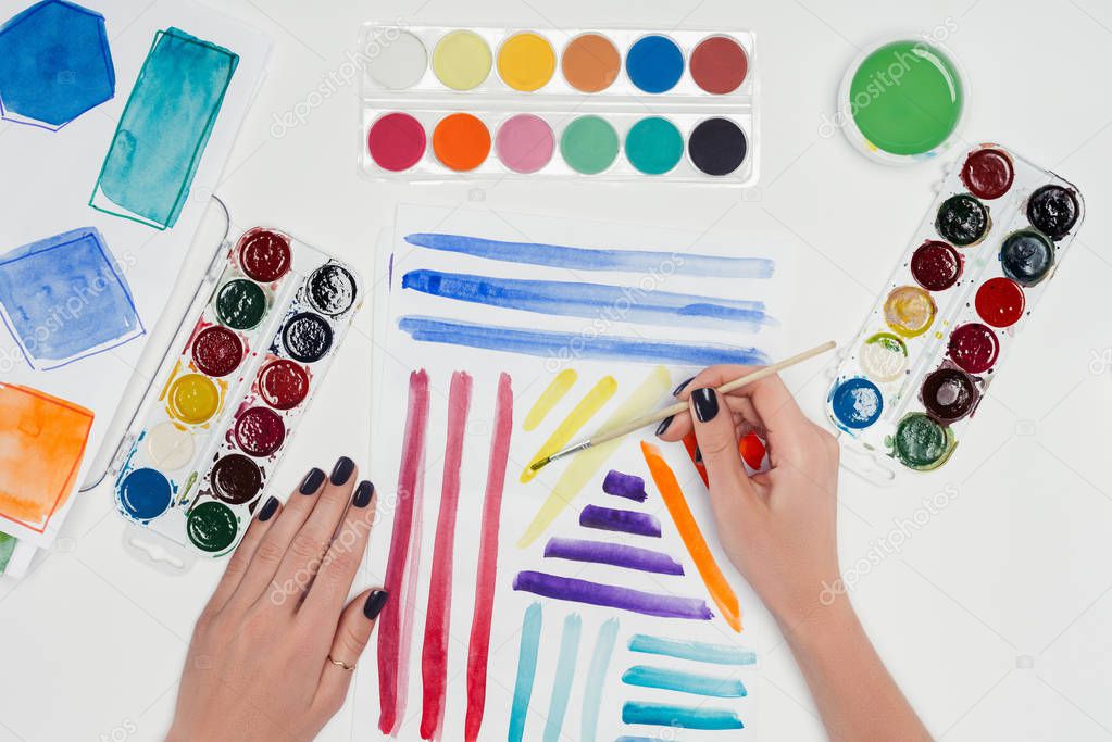 cropped image of female painter drawing colorful lines by paintbrush in textbook at white table