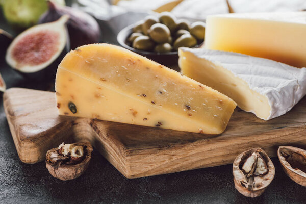 close up view of cheese assorted on wooden cutting board with hazelnuts and olives on dark surface