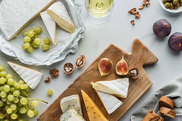 flat lay with food composition of cheese and fig pieces on cutting board, fruits and glass of wine on white marble surface