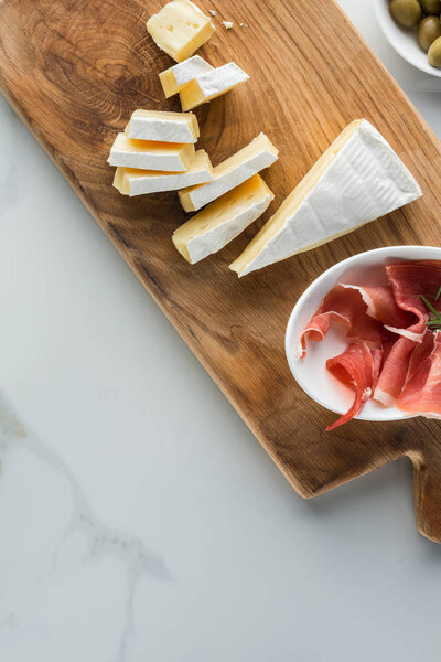 flat lay with camembert cheese and jamon on wooden cutting board on white marble tabletop